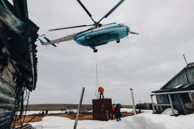 A helicopter delivering supplies to a remote snowy area with wooden cabins and two people.